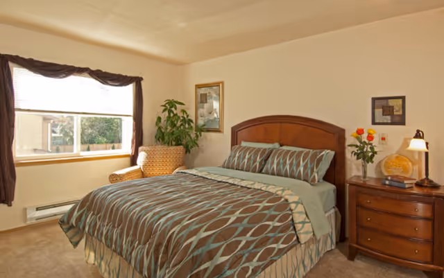 Furnished bedroom with a patterned bed, wooden headboard, nightstand with lamp and flowers, an armchair, and a window.