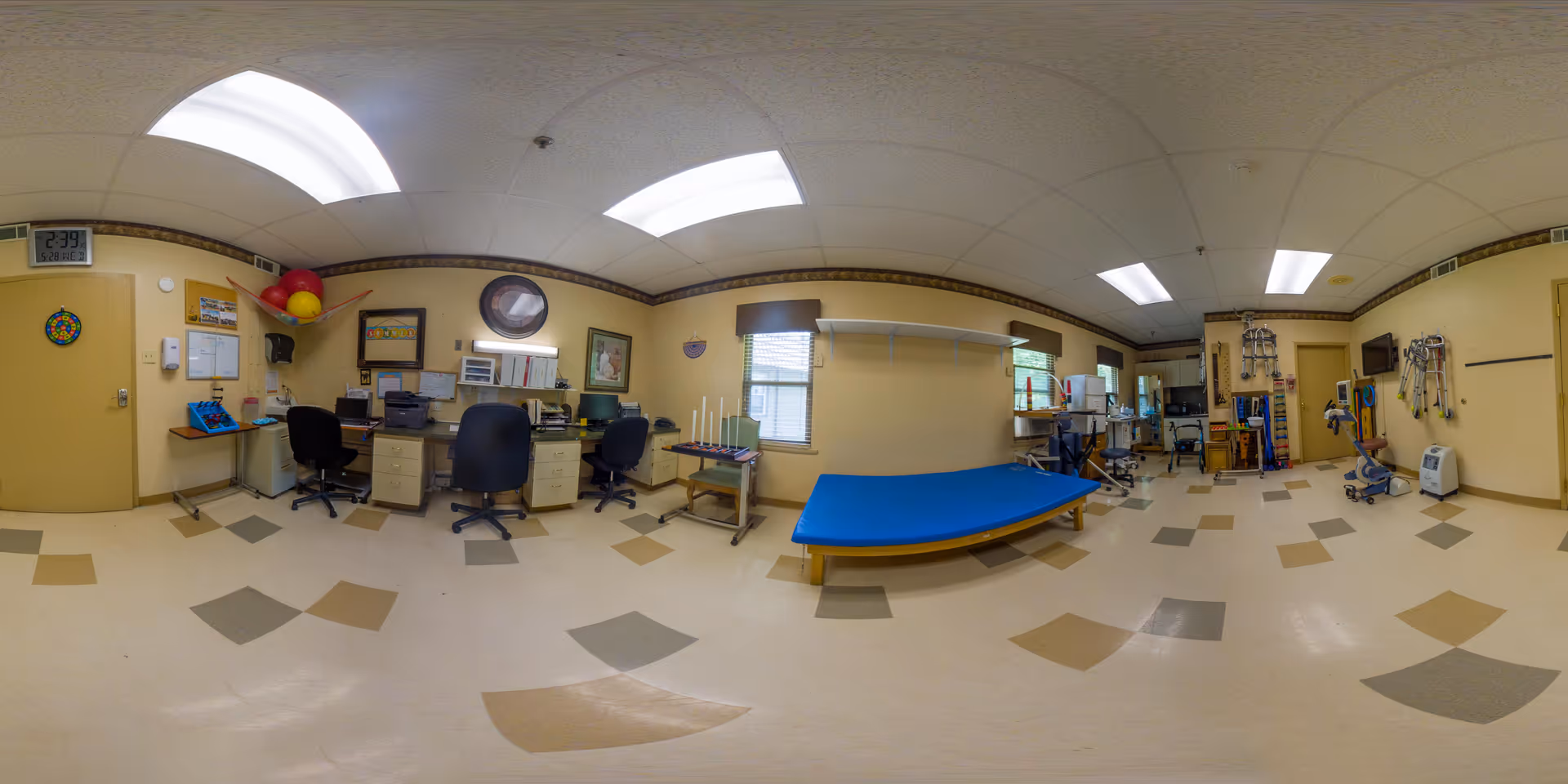 A spacious therapy or rehabilitation room with beige walls and a tiled floor featuring various colored square patterns. The room contains multiple office chairs and desks with computers and office supplies on the left side. In the center, there is a blue padded therapy table. The right side has various medical and therapy equipment, including walkers and exercise tools. The room is well-lit with fluorescent ceiling lights and has windows with blinds.