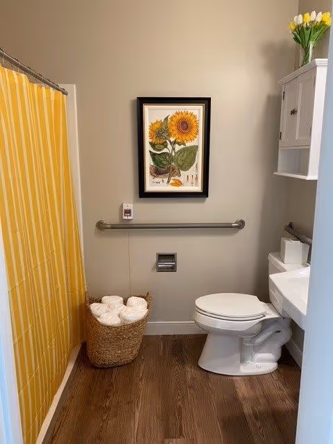 A bathroom with a yellow and white striped shower curtain on the left, a wicker basket filled with rolled white towels on the floor, a toilet with a white seat, a white cabinet mounted on the wall above the toilet, and a framed sunflower artwork hanging on the wall above a metal grab bar. The floor is wood-style and the walls are painted light beige.