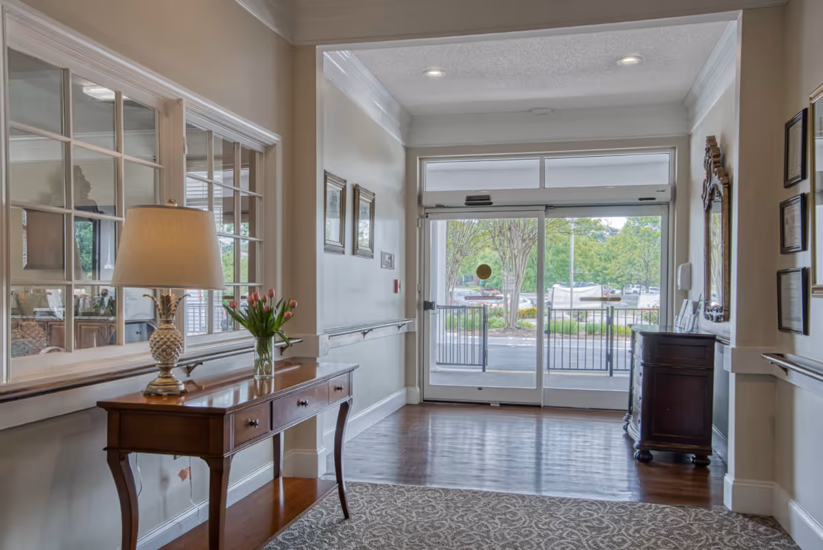 A bright entry lobby with a wooden console table holding a lamp and vase of flowers, patterned rug, and glass double doors leading outside.