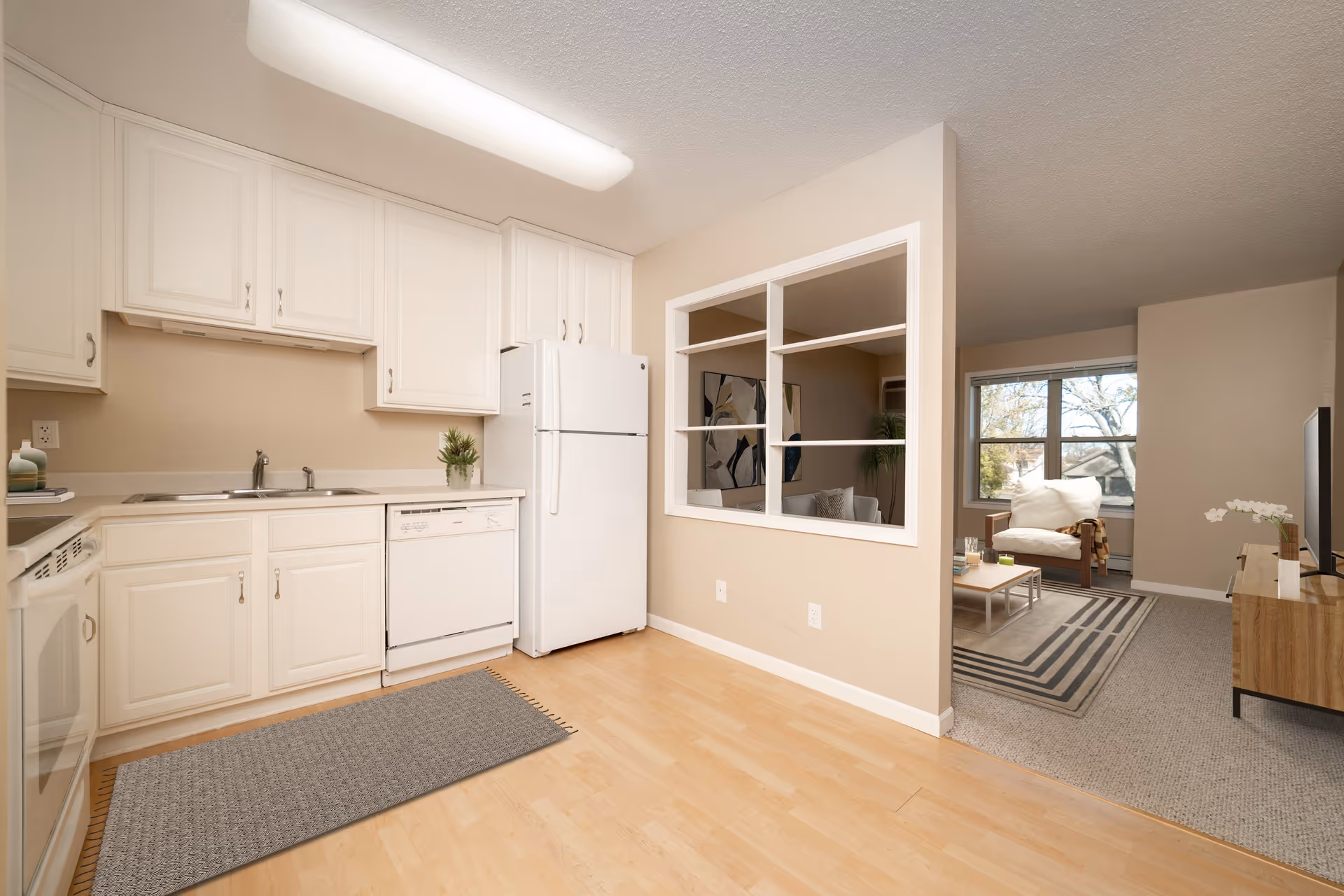 Interior view of a senior living facility kitchen with white cabinets, a white refrigerator, dishwasher, and sink. The kitchen has light wood flooring and a gray rug. Adjacent to the kitchen is a living room area with a large window, a beige armchair with a blanket, a coffee table, a TV on a wooden stand, and a decorative plant. A partial wall with open shelving separates the kitchen and living room.