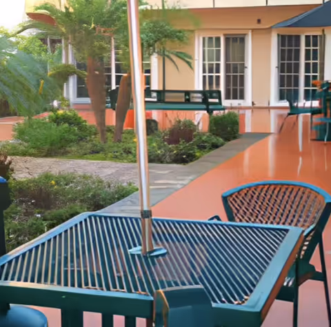 Outdoor patio area with metal tables and chairs, surrounded by greenery and plants. The patio is adjacent to a building with large windows and a peach-colored exterior. There is an umbrella pole in the center of the table in the foreground.