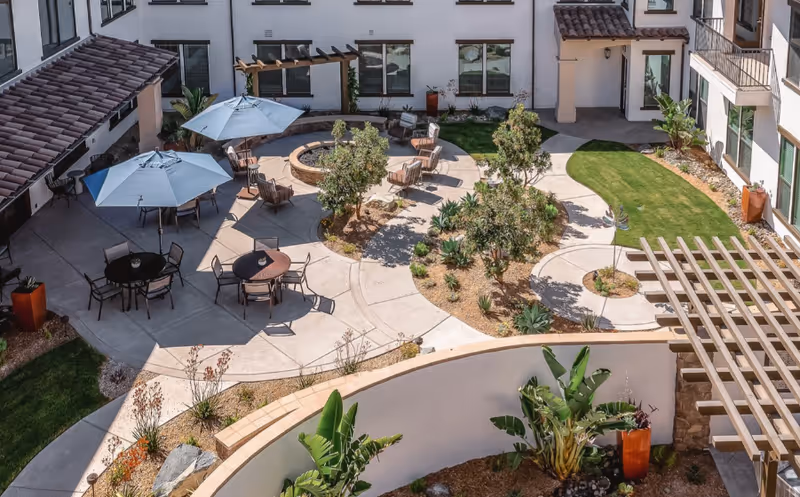 Aerial view of a landscaped courtyard with tables, umbrellas, chairs, a circular fire pit, walkways, and surrounding senior living building.