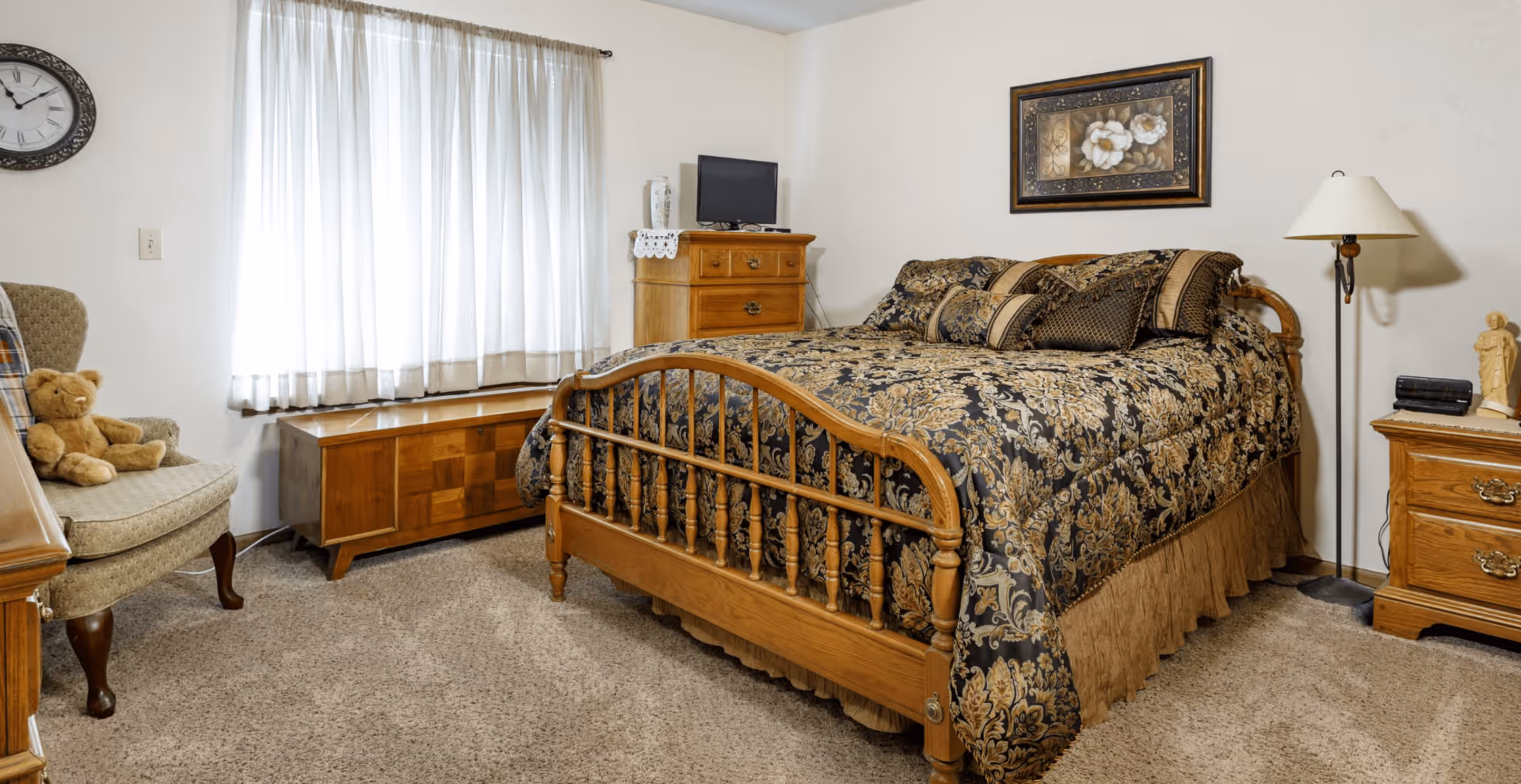 Traditionally furnished bedroom with a wooden bed dressed in patterned bedding, a dresser, armchair, lamp, and window with curtains.