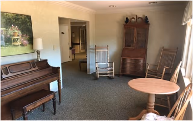 Cozy common room with an upright piano, two wooden rocking chairs, a round table and a wooden cabinet.