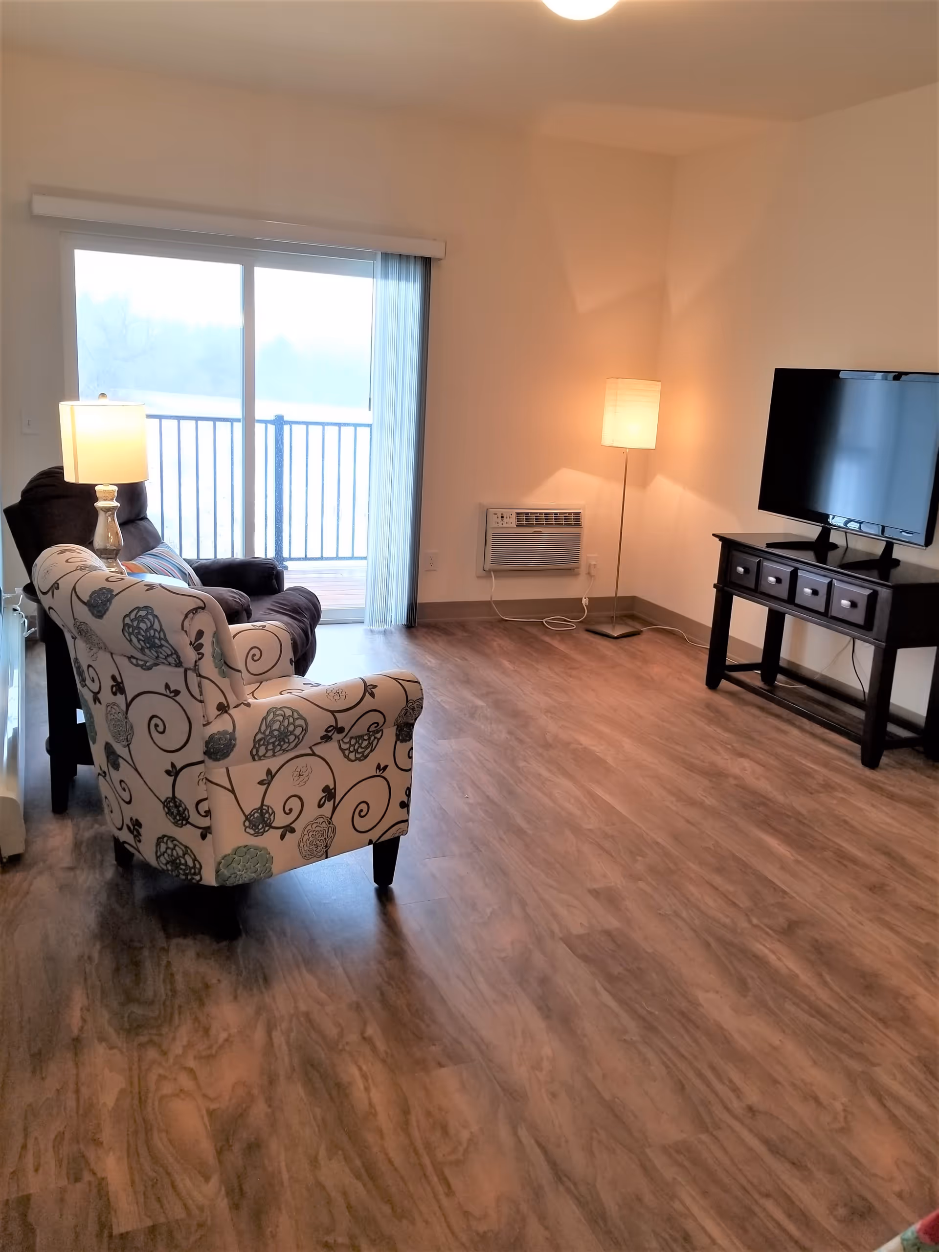 Bright living room with a patterned armchair and recliner, TV on a stand, floor lamp, and sliding glass door to a balcony.