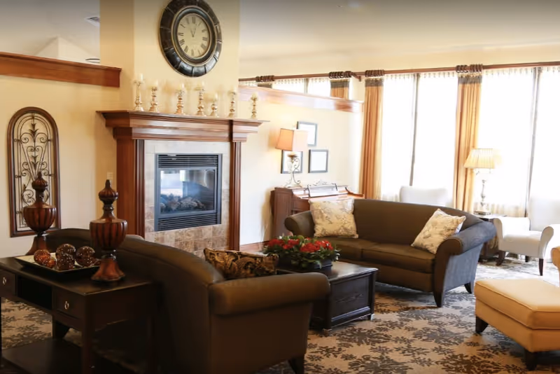 A cozy living room with two brown sofas, a wooden coffee table with a flower arrangement, a fireplace with a large clock above it, decorative candle holders on the mantel, and large windows with curtains allowing natural light to fill the room.