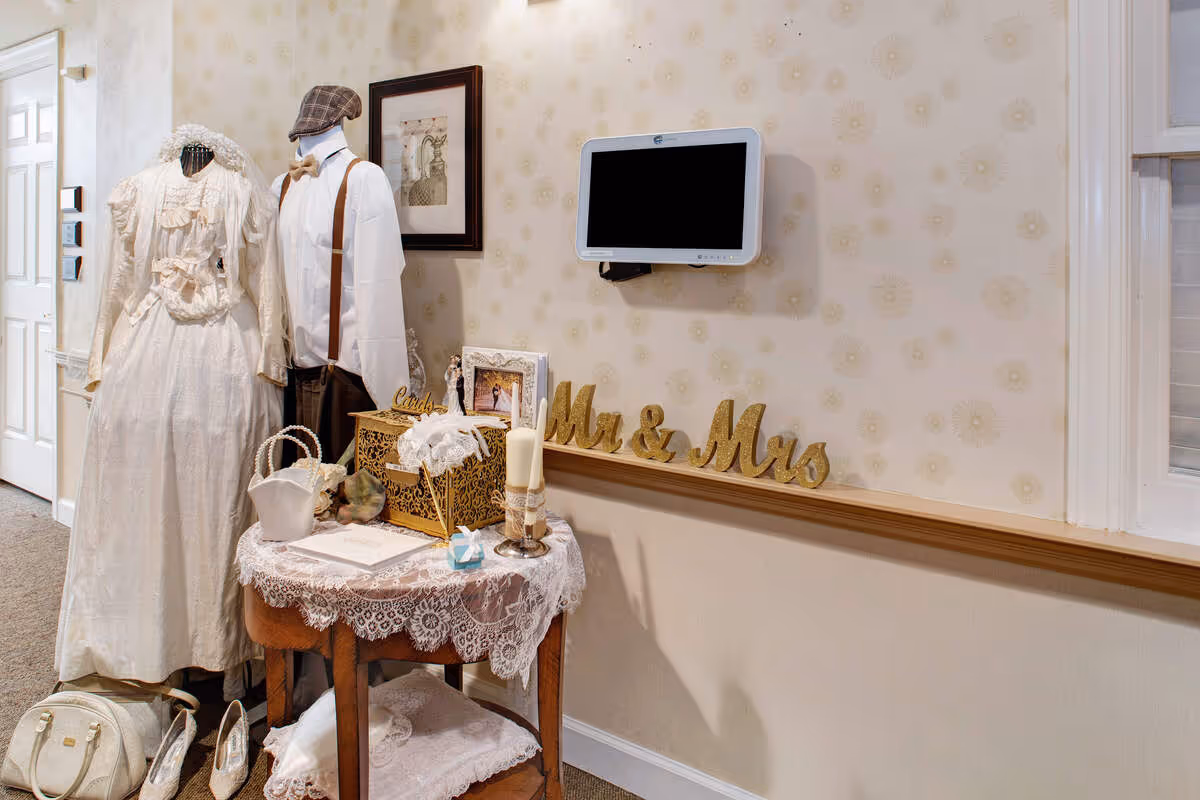 A corner of an interior room decorated with vintage wedding attire on two mannequins, one wearing a white lace wedding dress and the other dressed in a white shirt with suspenders and a flat cap. A small wooden table covered with a lace cloth holds wedding-themed items including a basket, a candle, a photo frame, and a decorative box. Gold glitter letters spelling 'Mr & Mrs' are displayed on a ledge against a wallpapered wall with a subtle floral pattern. A small wall-mounted monitor is also visible above the ledge.
