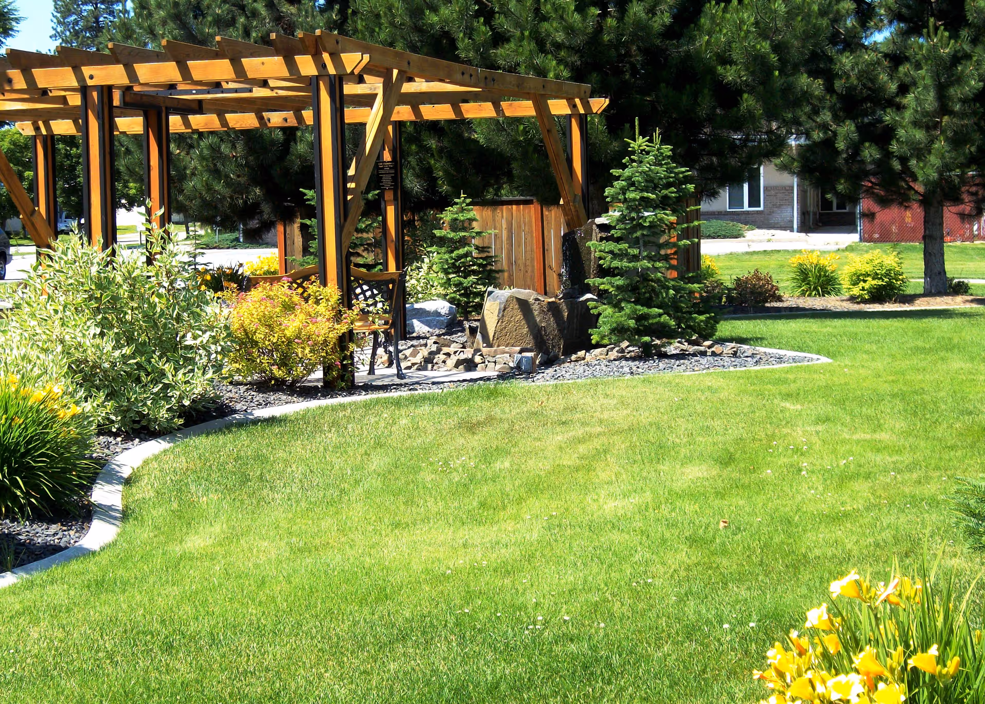 A well-maintained outdoor garden area with a wooden pergola, green grass, various shrubs, small pine trees, and yellow flowers. There is a stone water feature under the pergola and a building visible in the background.