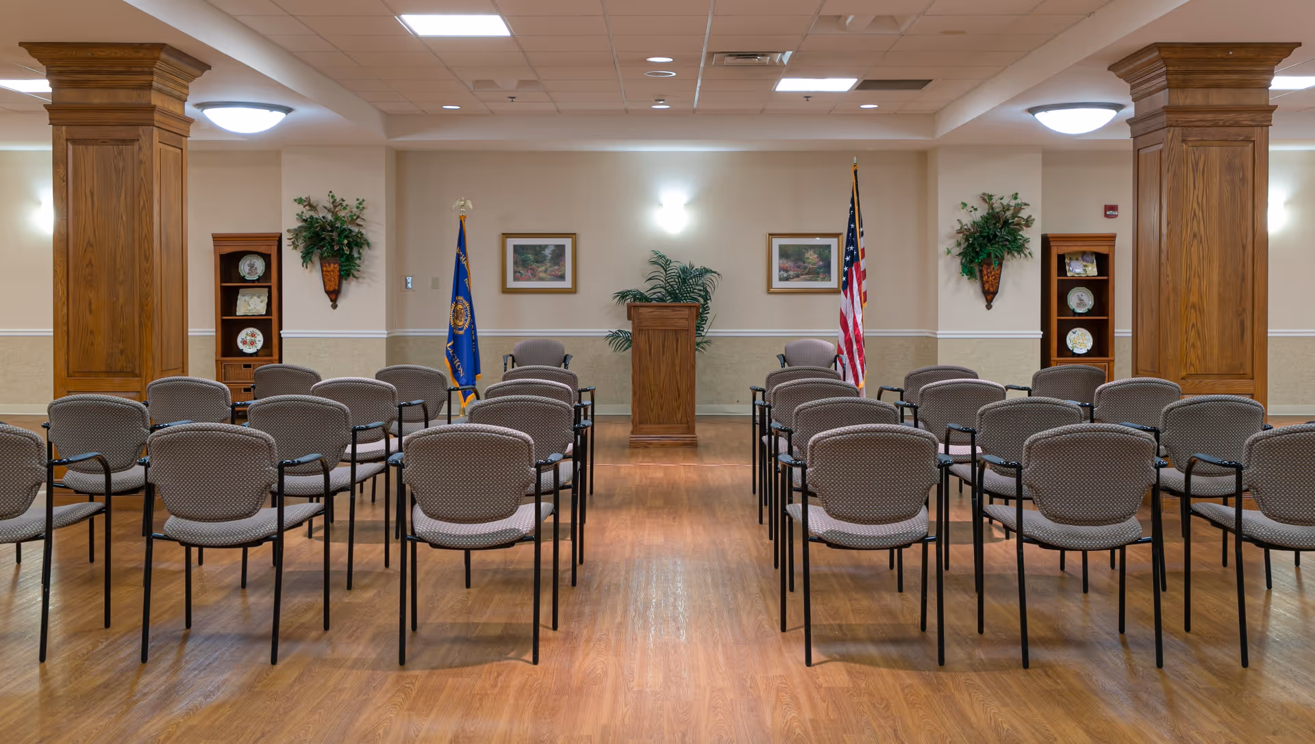 A meeting or conference room with rows of chairs facing a wooden podium. The room has wooden flooring, two large wooden pillars, two flags behind the podium, framed pictures on the wall, and decorative plants.