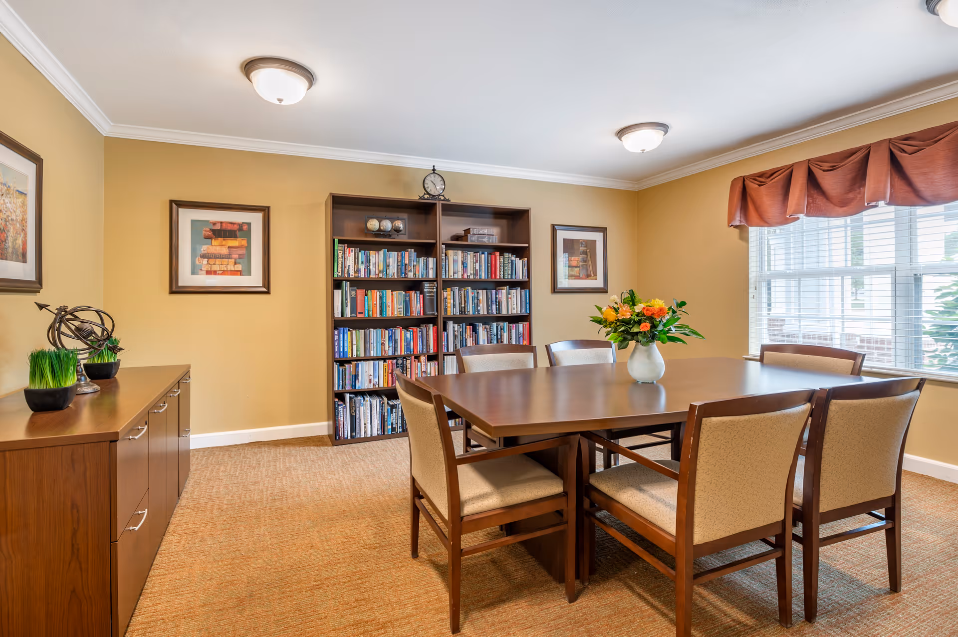 A well-lit communal dining room with a wooden table and chairs, a bookcase, sideboard, and a vase of flowers by a large window.
