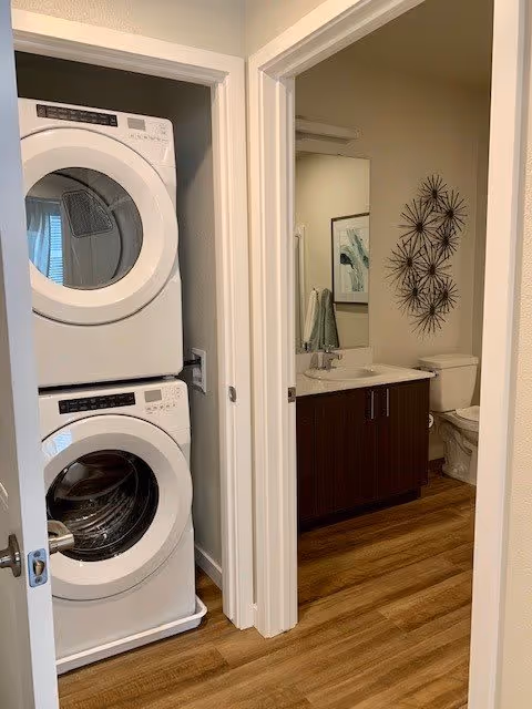 A small laundry area with a stacked washer and dryer unit on the left, next to a bathroom with a wooden vanity, sink, mirror, toilet, and decorative wall art. The floor is wood-style laminate.