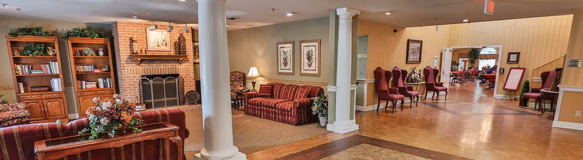 A spacious senior living facility common area with comfortable seating including striped sofas and wingback chairs. There is a brick fireplace with a decorative screen, wooden bookshelves filled with books and plants, framed botanical artwork on the walls, and a wooden coffee table with a floral arrangement. The area has columns, warm lighting, and a polished floor leading to another room with more seating.