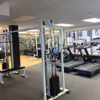 Interior view of a fitness room with exercise equipment including treadmills, weight machines, and benches. Large windows allow natural light to enter the space.