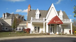 Exterior view of a senior living facility named Hearth & Home At Van Wert, featuring a large building with a peaked roof, white siding, and a red awning over the entrance. The sky is clear with a few clouds, and there is a driveway and some landscaping with trees and bushes in front.