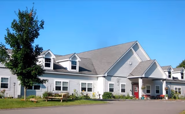 Exterior view of a single-story senior living facility building with gray siding, multiple windows, a pitched roof with dormer windows, a red entrance door, and a small tree and bench on the lawn in front under a clear blue sky.