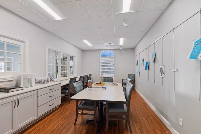 A bright room with a long table surrounded by chairs, wood flooring, and a wall with artwork hanging on display wires. There are windows on one side and a cabinet with drawers on the other side.