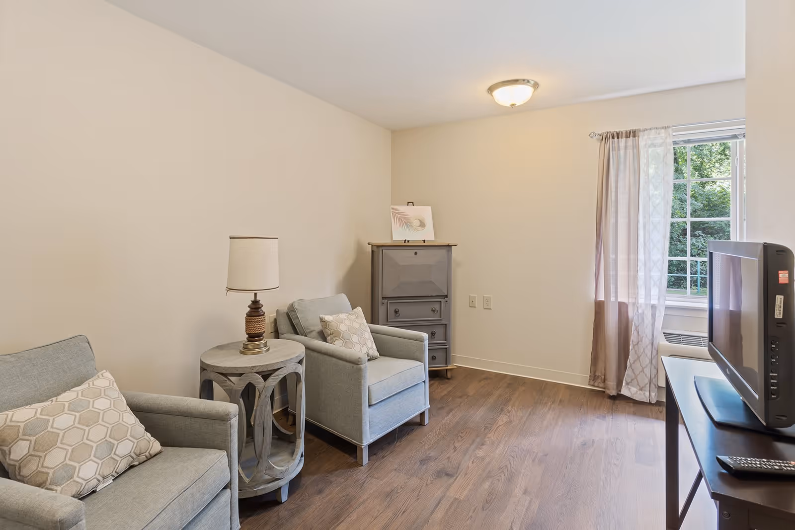 A small living room area with two light gray armchairs, each with a patterned cushion. Between the chairs is a round wooden side table with a lamp on it. In the corner is a gray cabinet with a decorative picture on top. A window with sheer and solid curtains lets in natural light. A flat-screen TV sits on a dark wooden table with a remote control in front of it. The room has wooden flooring and beige walls.