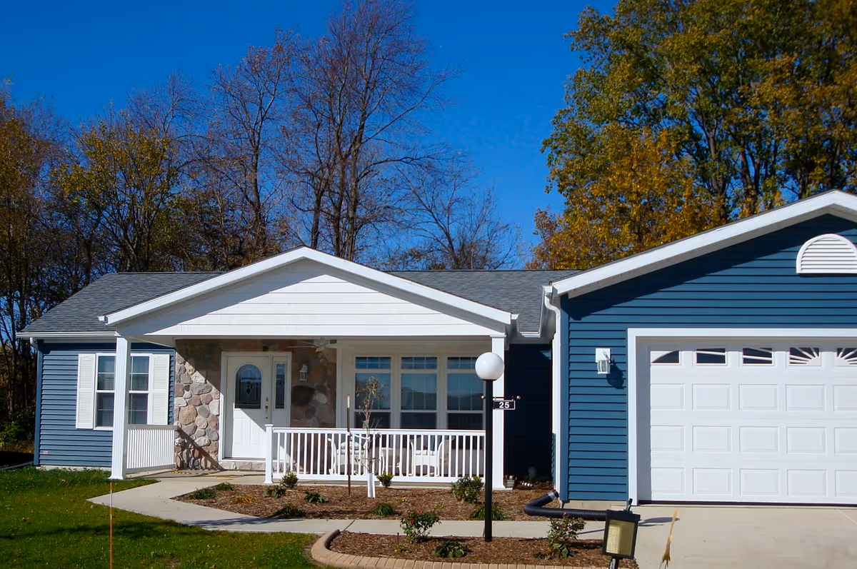 Single-story house with blue siding and white trim, featuring a front porch with white railing, a stone accent wall around the front door, a two-car garage, and a small landscaped area with shrubs and a lamp post in front. Trees with autumn foliage are visible in the background under a clear blue sky.