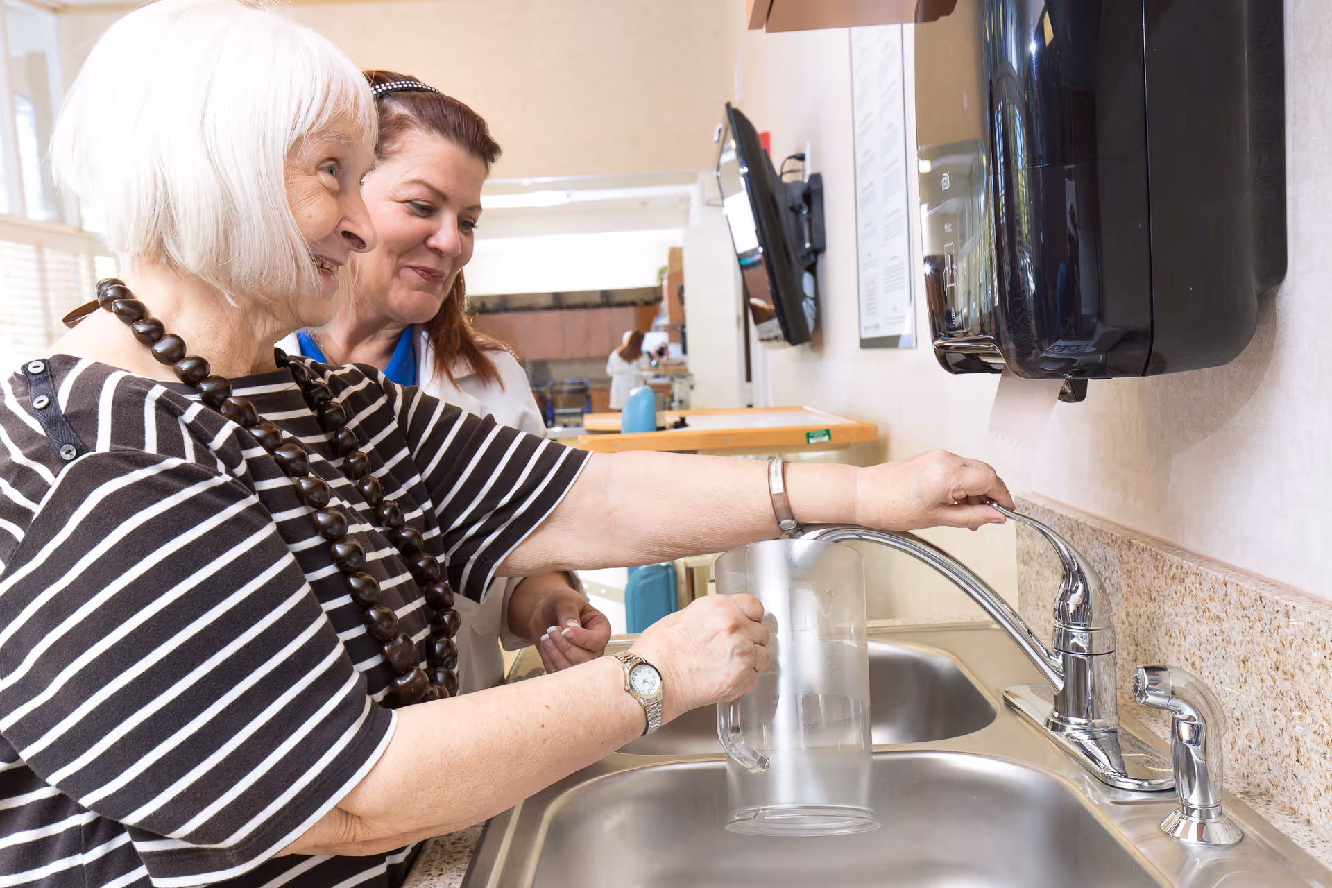 An elderly woman with white hair and a striped shirt is filling a clear pitcher with water from a kitchen sink faucet, while a caregiver stands behind her smiling and assisting.