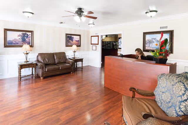 A reception area in a senior living facility with a wooden reception desk where a woman is seated and talking on the phone. The room has wooden flooring, a brown leather couch with two side tables and lamps, framed landscape paintings on the walls, and a floral arrangement on the desk.