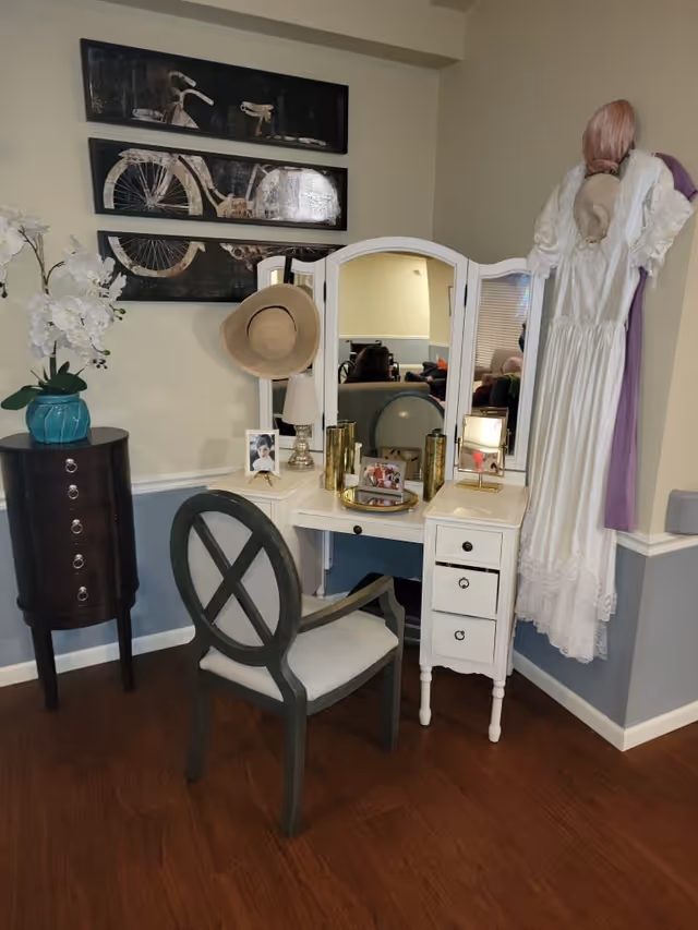 A cozy corner with a white vanity table featuring a tri-fold mirror, a gray cushioned chair, and decorative items including framed photos, a lamp, and gold vases. A wide-brimmed hat hangs on the mirror. To the right, a white vintage dress with a pink headscarf is displayed on the wall. On the left, a small dark wooden chest with drawers holds a blue vase with white flowers. Above the chest, three black panels with a bicycle design are mounted on the wall. The room has wood flooring and light blue and beige walls.