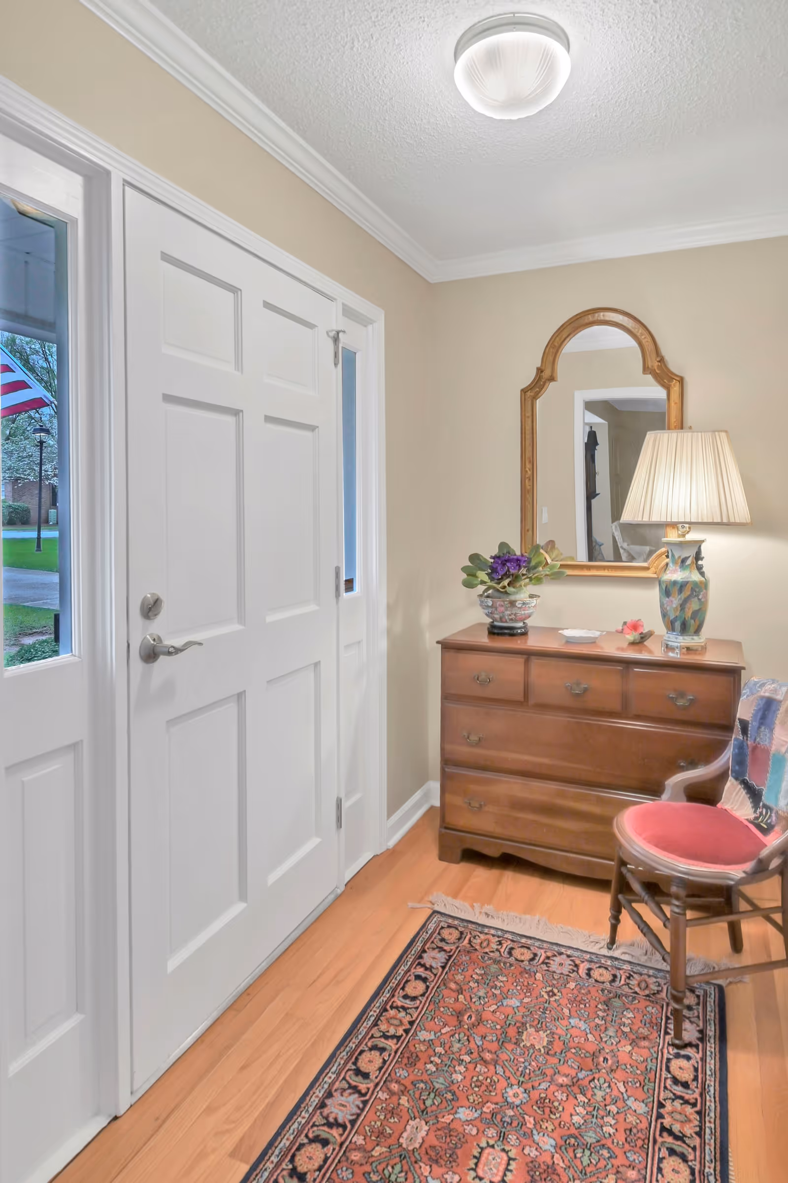 Entryway with a white front door and sidelights, a wooden chest of drawers with a decorative lamp and a potted plant on top, an ornate mirror hanging above the chest, a wooden chair with a red cushion and a colorful throw, and a patterned rug on the wooden floor.