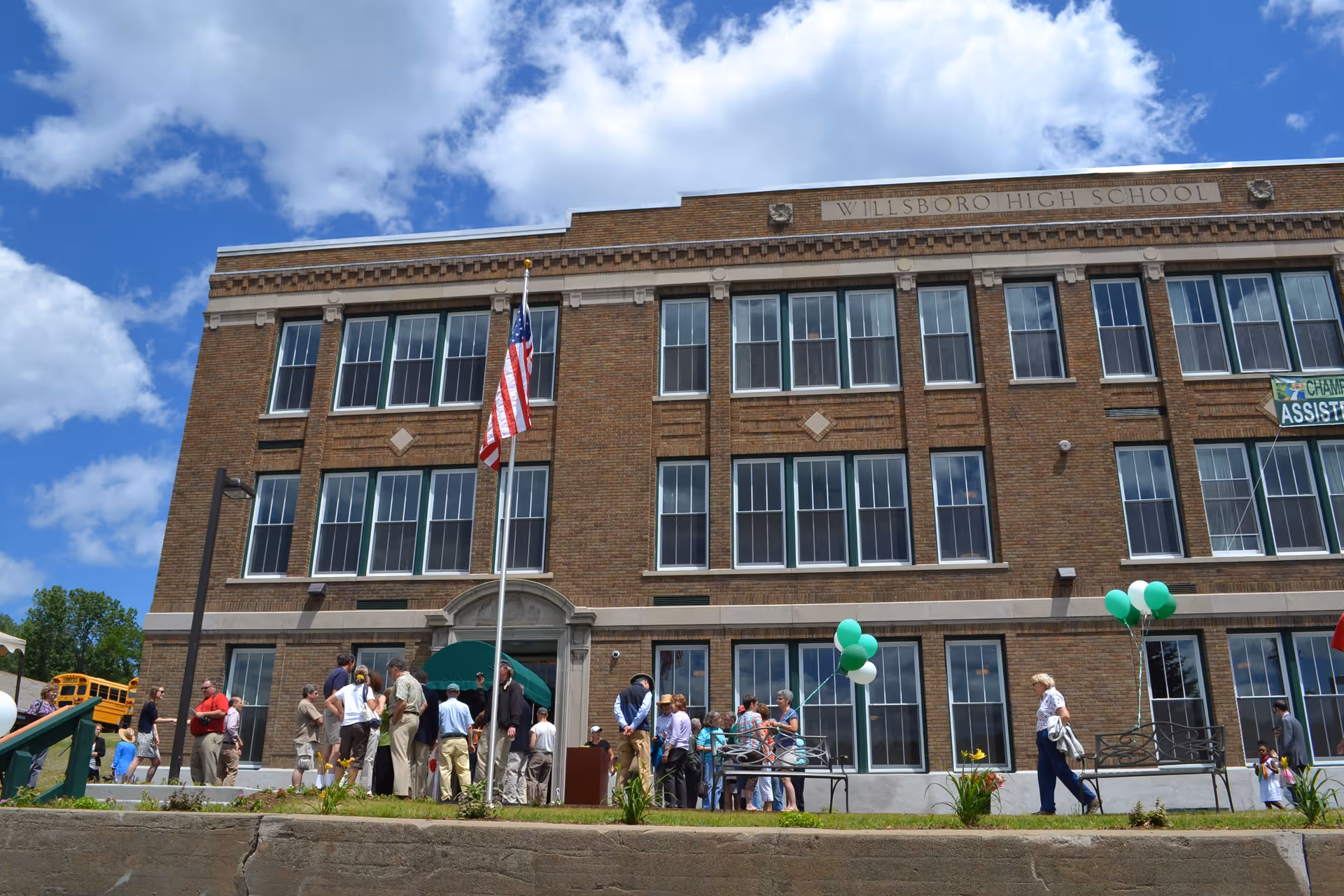 A group of people gathered outside a large brick building labeled Willsboro High School under a partly cloudy blue sky. There are green and white balloons tied to benches and an American flag on a flagpole in front of the building.