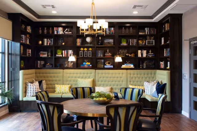 A cozy seating area with a round wooden table surrounded by six striped chairs. Behind the table is a built-in green cushioned bench with various patterned throw pillows. The back wall features dark wooden bookshelves filled with books, decorative items, and small plants. Two pendant lights hang above the bench, and a large window is visible on the left side, letting in natural light.