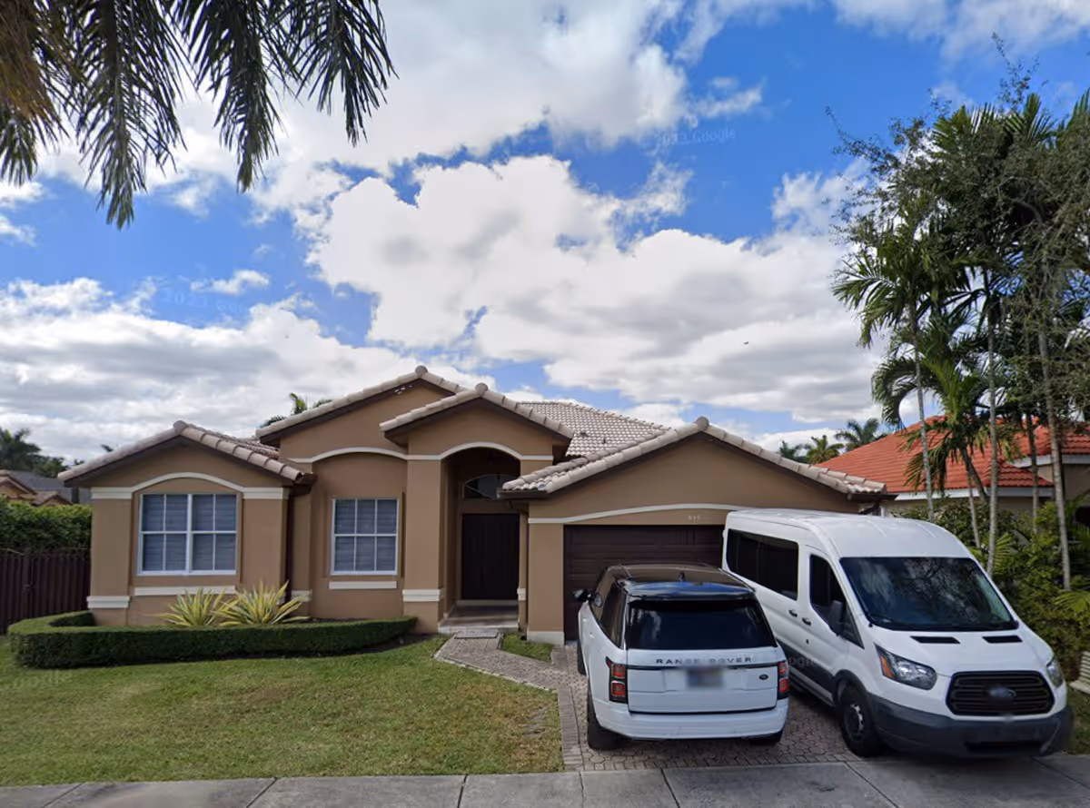 Front exterior view of a single-story beige house with a tiled roof, two vehicles parked in the driveway, and palm trees around the property under a partly cloudy sky.