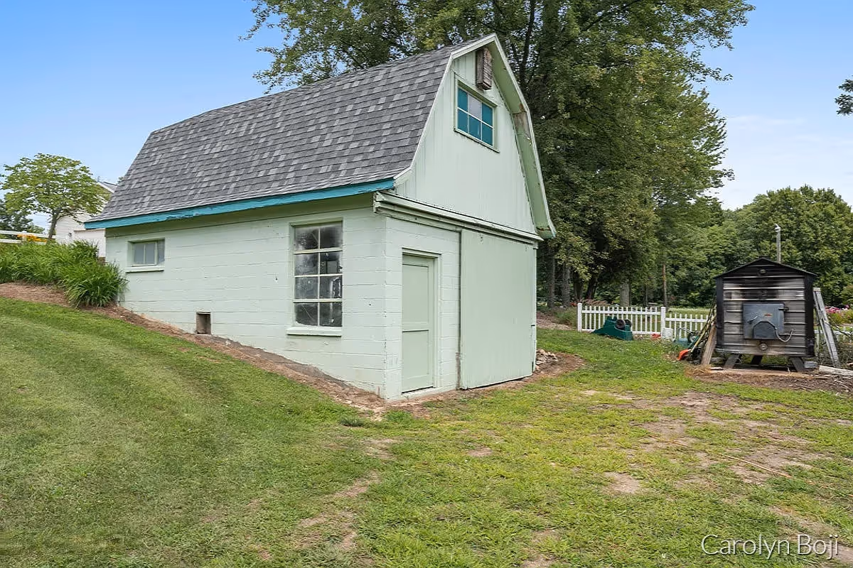 Small pale green two-story outbuilding with a sloped roof on a grassy yard beside trees and a smaller shed.
