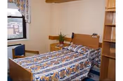 A small bedroom with a single bed covered in a patterned bedspread. The bed has a wooden headboard and footboard. Next to the bed is a wooden nightstand with a small plant and a telephone. There is a window with floral curtains on the left wall and a wooden shelving unit on the right side of the room.