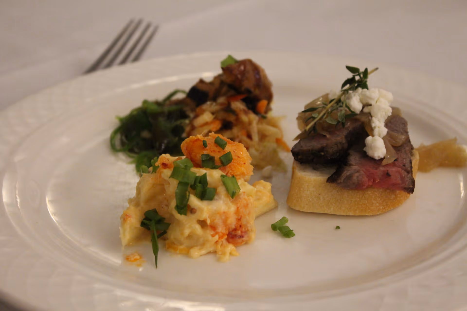 A white plate with three different appetizers: a creamy seafood salad garnished with chopped green onions, a small portion of mixed greens and vegetables, and a slice of bread topped with slices of rare steak, caramelized onions, goat cheese, and a sprig of thyme.
