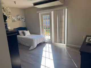 A bright bedroom with a single bed covered in white bedding, a dark dresser with decorative items, and a glass door with vertical blinds allowing sunlight to stream into the room.