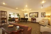 A cozy living room area in Garden View Post-Acute Rehab featuring a grand piano, several armchairs, a sofa, a wooden side table with books, framed artwork on the walls, and large windows letting in natural light.