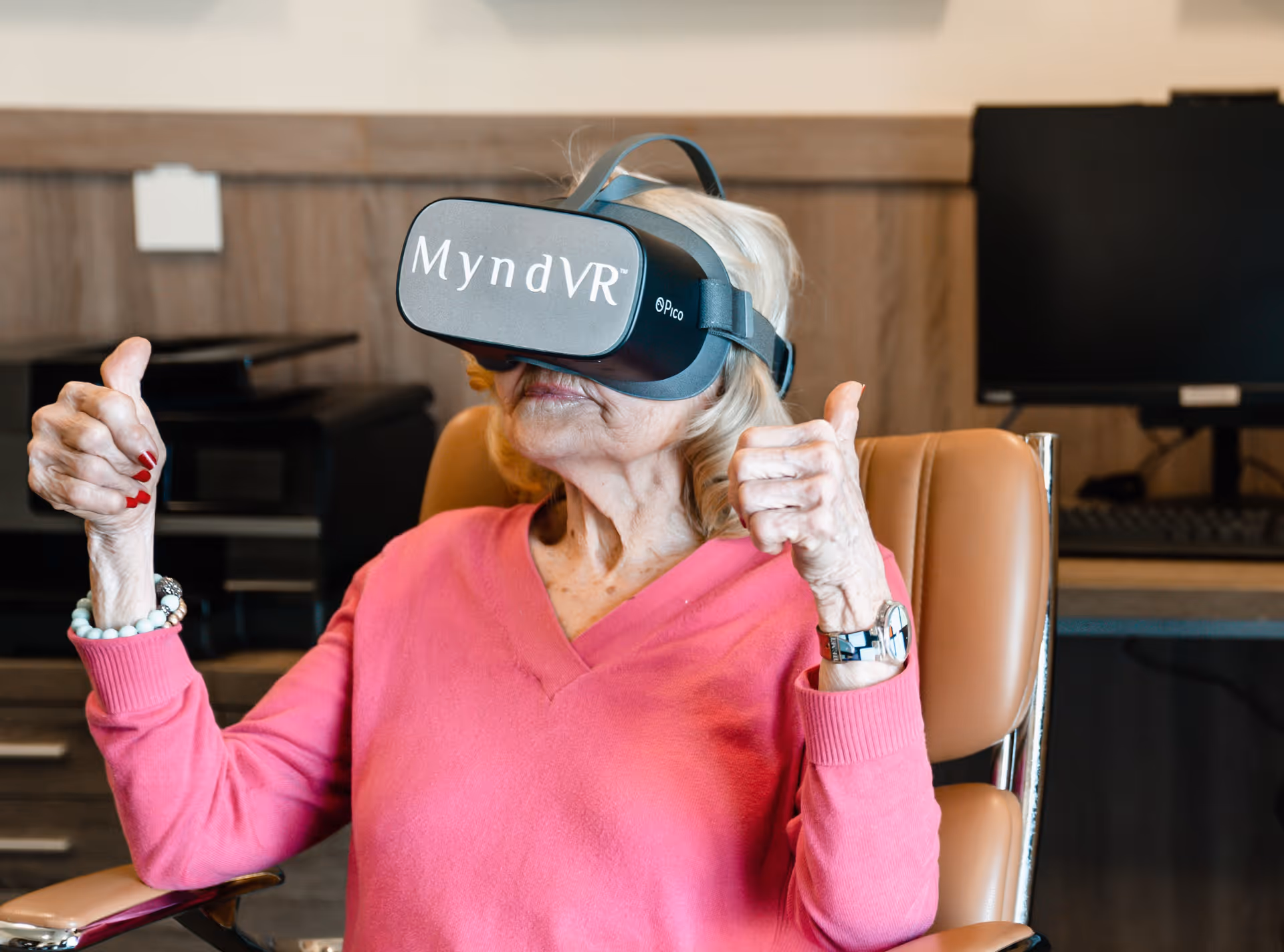 An elderly woman wearing a pink sweater is sitting in a chair and using a MyndVR virtual reality headset, giving two thumbs up. The background shows a desk with a computer monitor and keyboard.