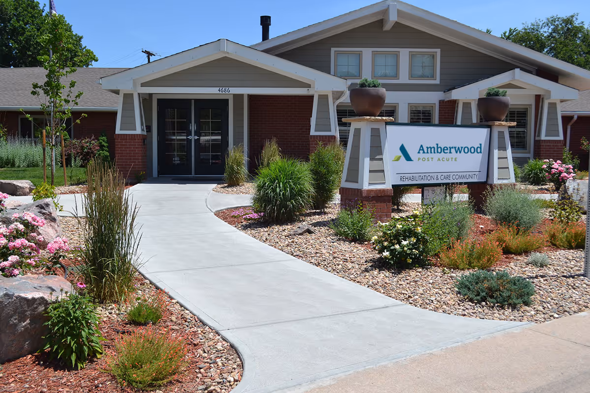Front exterior view of Amberwood Post Acute rehabilitation and care community building with a concrete walkway, landscaped garden with rocks, bushes, and flowers, and a sign displaying the facility name.
