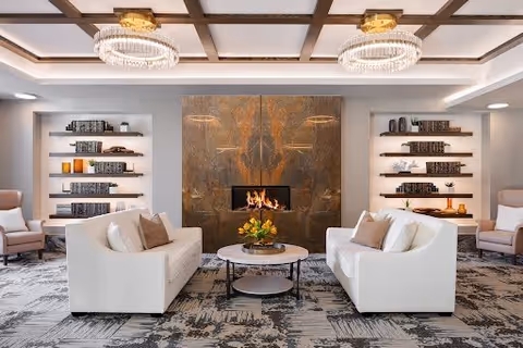 A modern living room with two white sofas facing each other across a round coffee table with a flower arrangement. Behind the table is a fireplace set in a large stone wall. On either side of the fireplace are built-in shelves with books and decorative items. The ceiling features wooden beams and two elegant chandeliers. The room has a patterned carpet and two armchairs near the walls.