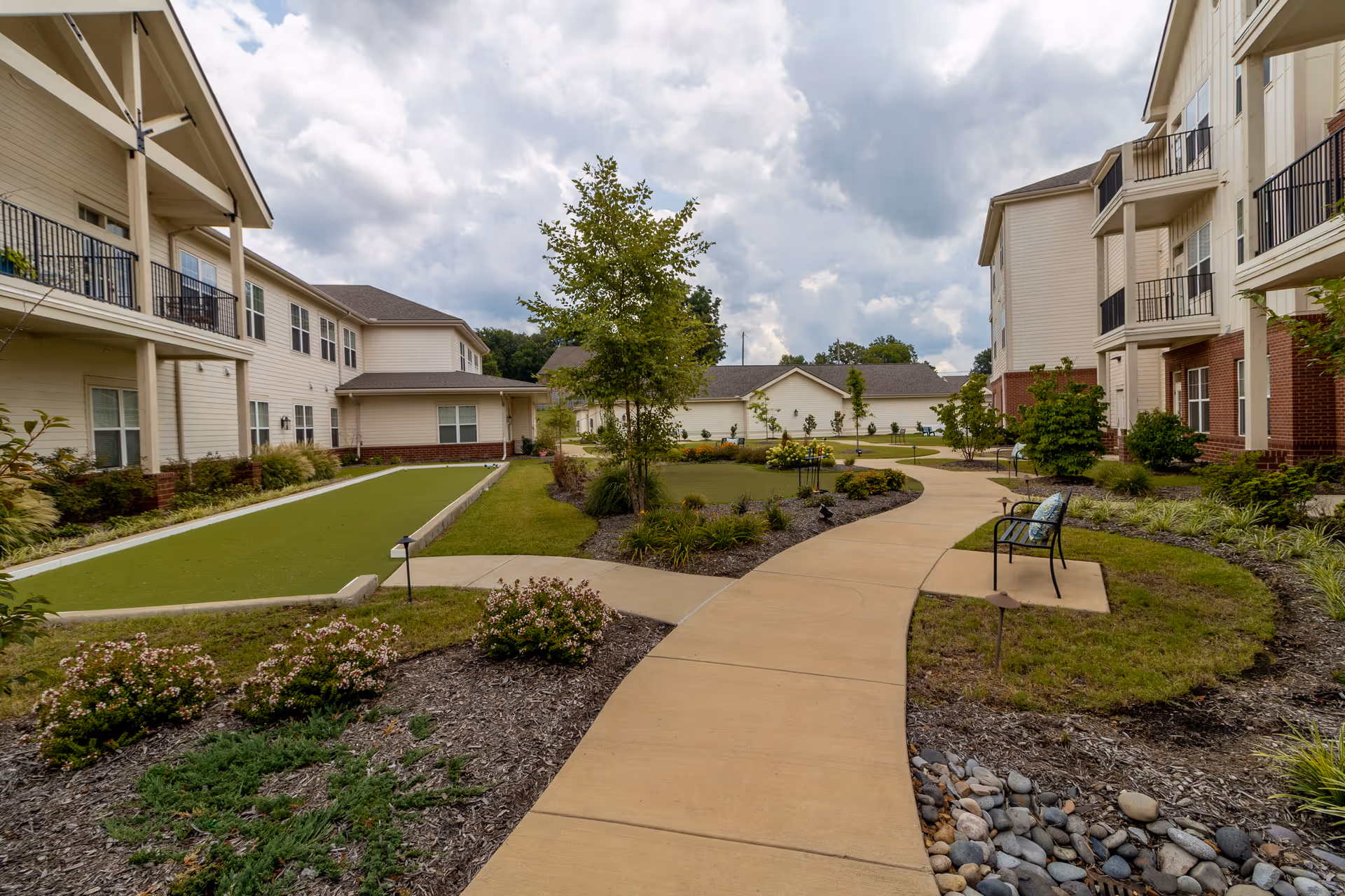 Outdoor courtyard area at StoryPoint Cordova featuring a winding concrete pathway, landscaped garden beds with shrubs and flowers, a small tree in the center, benches with cushions, and two-story residential buildings with balconies on either side under a cloudy sky.
