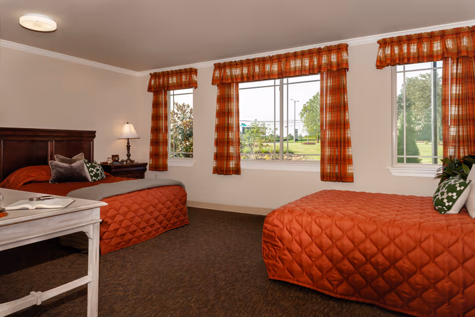 A bright assisted living bedroom with two twin beds covered in rust-colored quilts, a wooden headboard, nightstand with a lamp, and three windows with plaid curtains overlooking greenery.