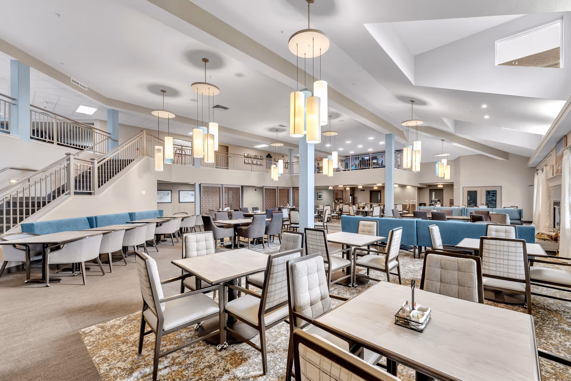 Spacious dining area with multiple tables and chairs arranged neatly. The room features modern pendant lighting hanging from a high ceiling, blue cushioned bench seating along the walls, and a staircase leading to an upper level with railings. The space is well-lit with natural and artificial light, creating a welcoming atmosphere.