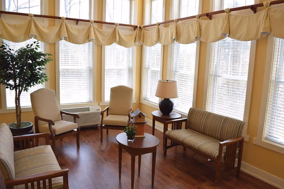 A bright sitting area with multiple windows covered by beige valances. The room features wooden flooring, two beige armchairs, two striped cushioned wooden sofas, a small round wooden table with a lamp, and a smaller oval wooden table with a small plant and a decorative birdhouse. There is also a potted plant in the corner.