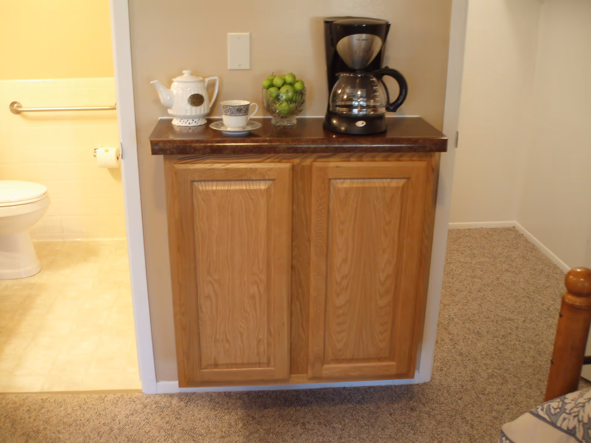 A small wooden cabinet with a dark countertop holding a coffee maker, a white teapot, a teacup with saucer, and a glass bowl filled with green apples. To the left, there is a partial view of a bathroom with a toilet and a grab bar on the wall. The floor is carpeted outside the bathroom and tiled inside.