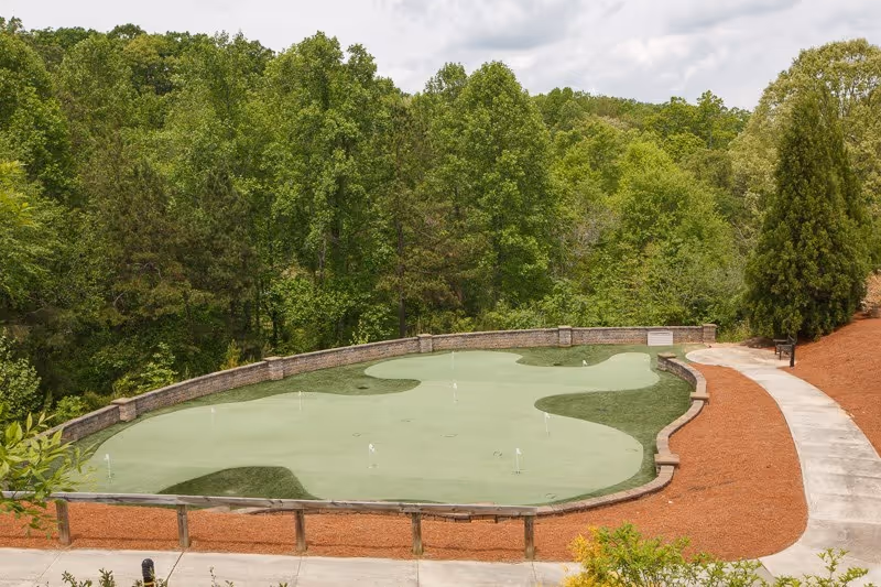 Outdoor putting green surrounded by a low stone wall and a curved concrete pathway, with dense green trees in the background under a cloudy sky.