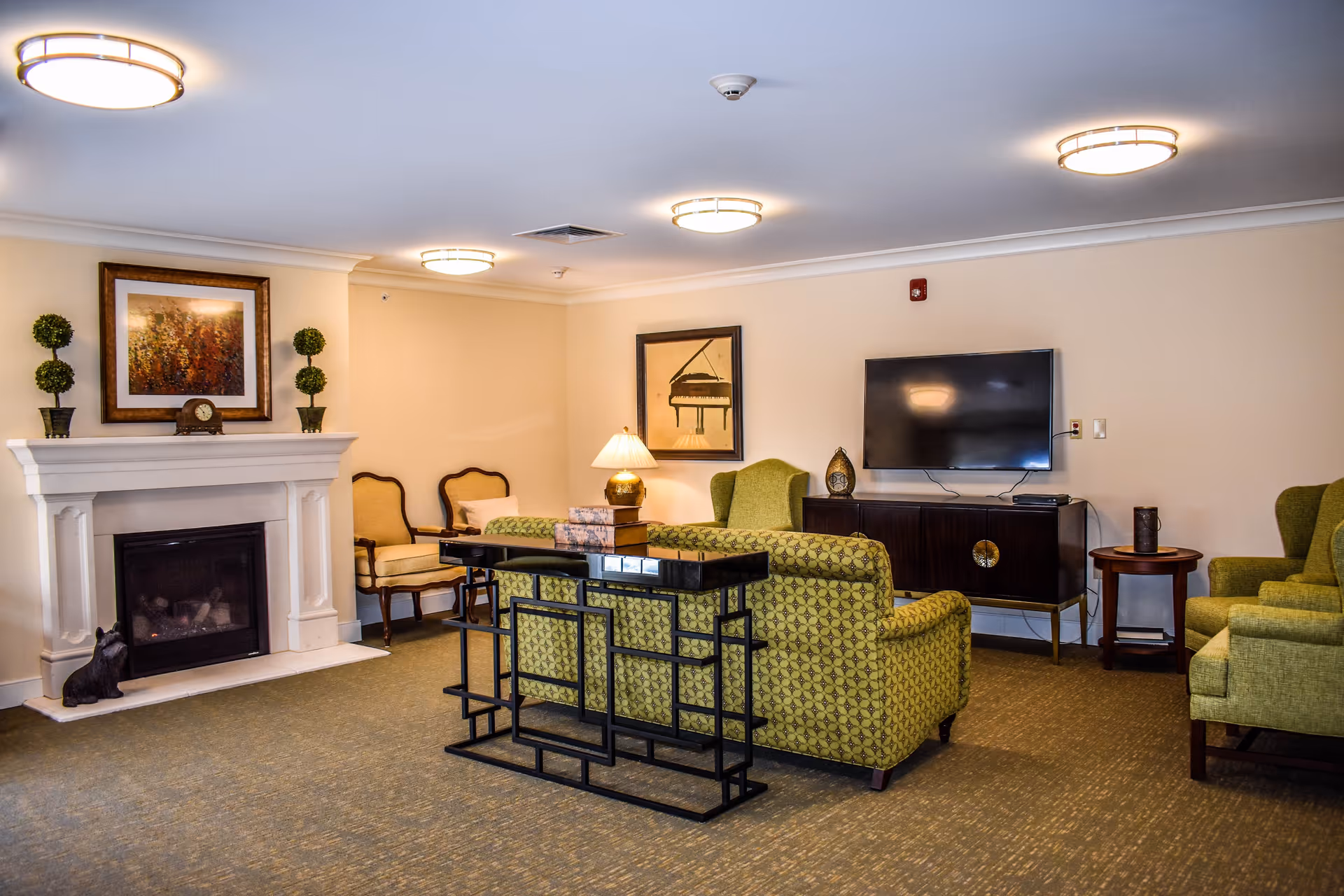 A cozy living room in a senior care facility featuring green patterned sofas and armchairs, a white fireplace with decorative plants and a framed picture above it, a flat-screen TV mounted on the wall, a dark wooden cabinet, and warm lighting from ceiling fixtures and a table lamp.