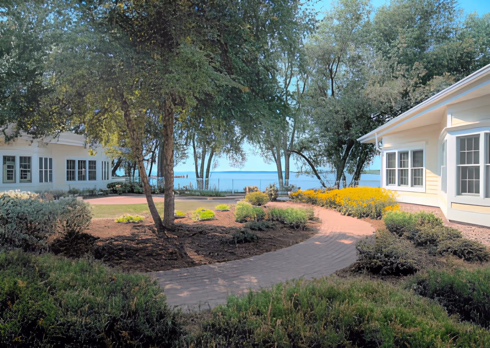 A landscaped outdoor garden area with a paved walkway winding through bushes, trees, and yellow flowers. Two light-colored buildings with multiple windows are visible on either side, and a body of water can be seen in the background beyond the trees under a clear blue sky.