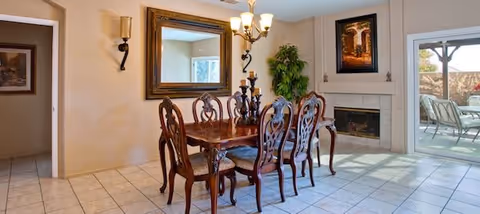 A dining room with a wooden dining table and six ornate chairs. The room features a large mirror on the wall, two wall sconces, a chandelier above the table, a fireplace with a framed picture above it, a potted plant in the corner, and a sliding glass door leading to an outdoor patio with chairs.