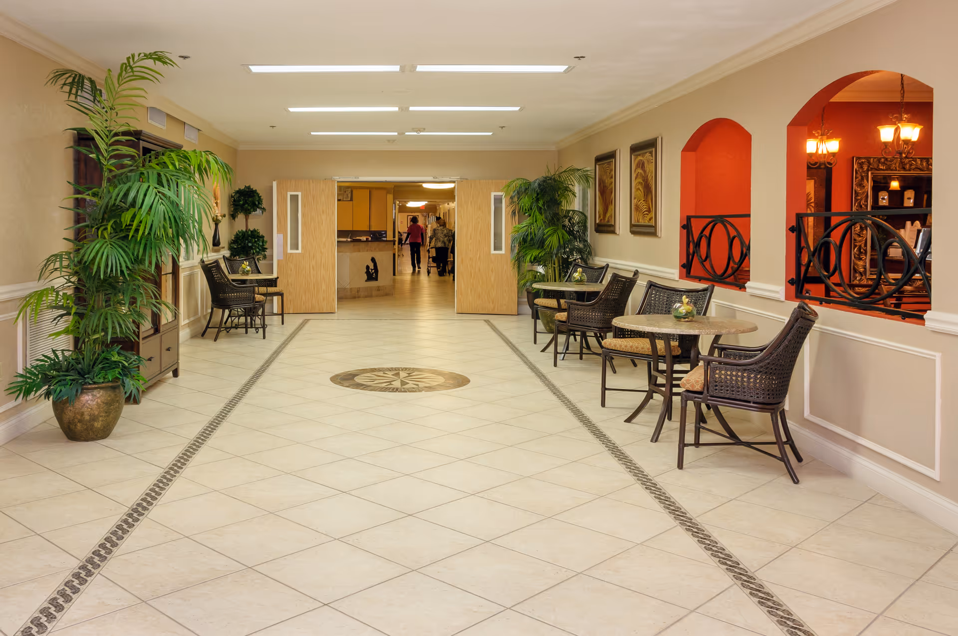 A spacious, well-lit hallway in a senior living facility with tiled floors and decorative inlay. The hallway features several small tables with wicker chairs along the right side, potted plants on both sides, framed artwork on the walls, and double wooden doors at the far end. Two people are visible walking in the distance through the open doors.