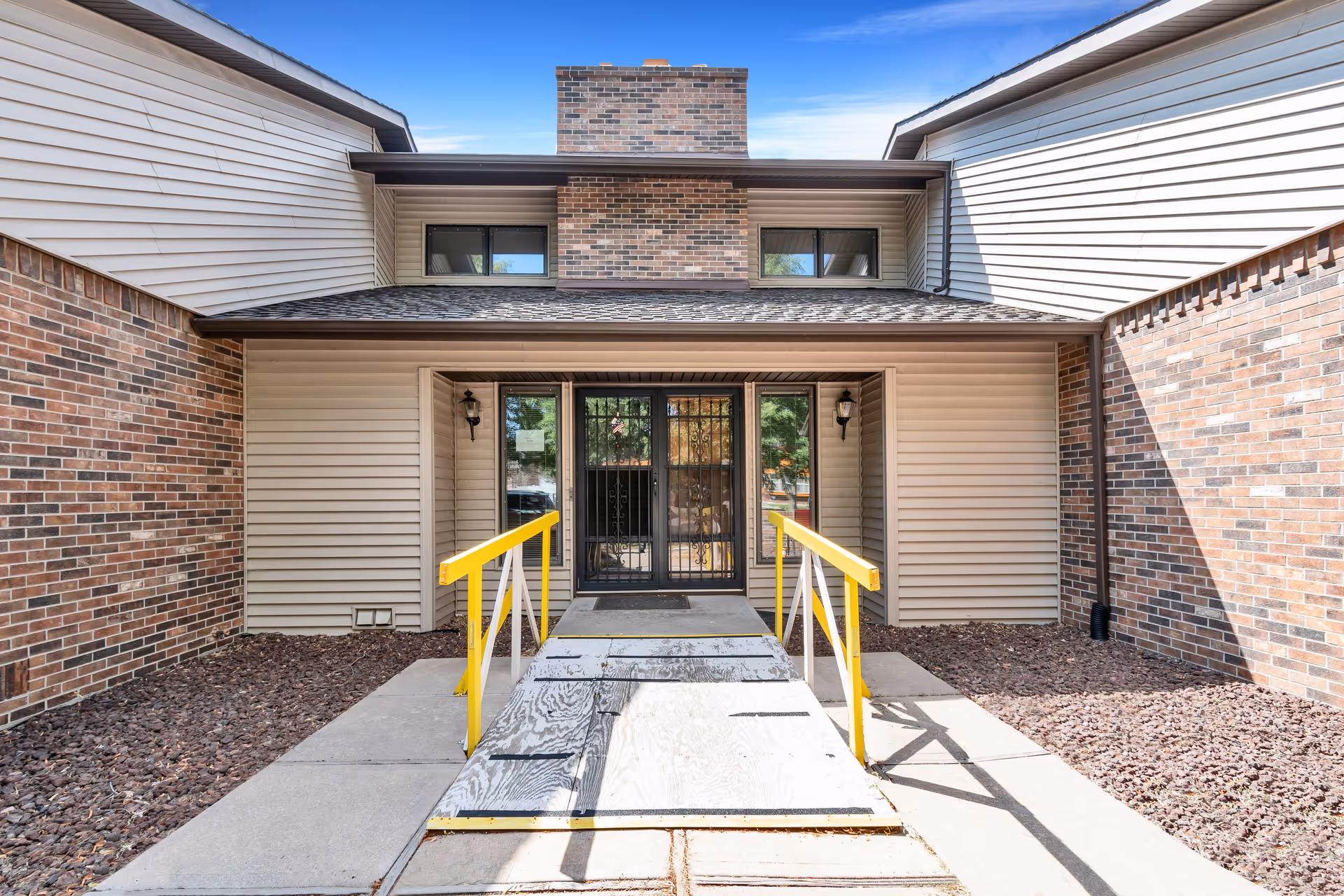 Entrance to a building with a wooden ramp featuring yellow handrails leading to double glass doors with metal security bars. The building exterior has beige siding and brick walls with a chimney above the entrance. Two wall-mounted lantern-style lights flank the doors.