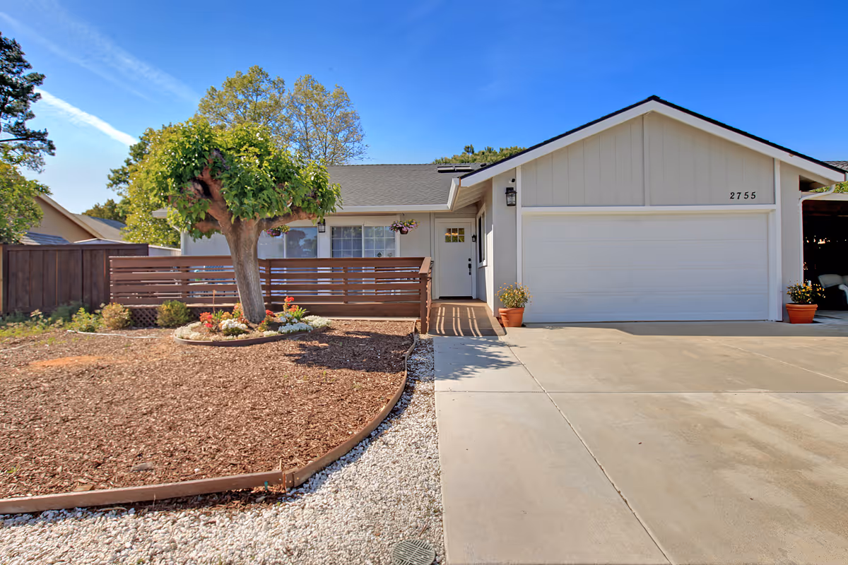 Front exterior view of a single-story house with a white garage door, a small porch with a white door, a tree in the mulched front yard, and a clear blue sky.